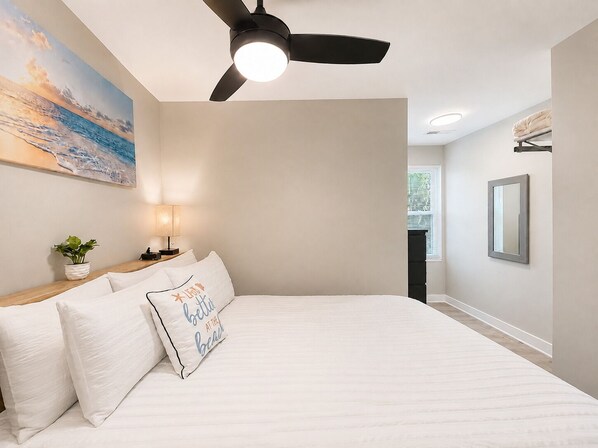 A bedroom with a bed, a ceiling fan, a nightstand with a lamp, a framed picture on the wall, and a window with blinds. - #101 Ocean Inn 2 Bedroom 1.5 Bath Modern Favorite (Isle of Palms)