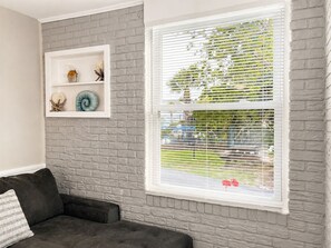 A room with a grey brick wall, a white-framed window with blinds, a dark grey sofa, and a shelf with decorative items.