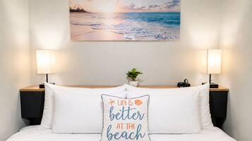 Quarto de hotel com uma cama, duas luminárias de cabeceira, uma mesinha de apoio, um quadro na parede com uma paisagem de praia e um travesseiro com uma frase sobre o mar.