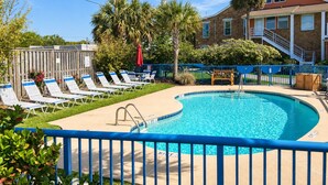 A swimming pool with lounge chairs, a wooden deck, and a building with a red roof in the background.