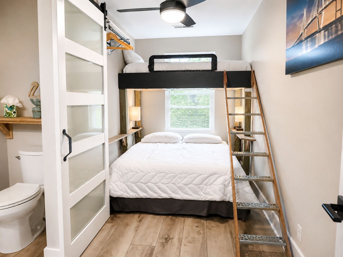 Un dormitorio pequeño con una cama nido, una escalera de madera, una mesita de noche con lámpara y una ventana con persianas.