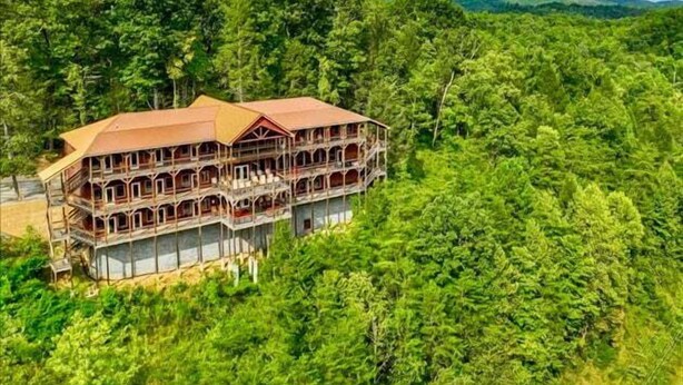 A large, multi-story building with a red roof, surrounded by dense forest. - Sycamore Mansion | 14 Suites | Arcade | Fire Pit | Amazing Views (Gatlinburg)