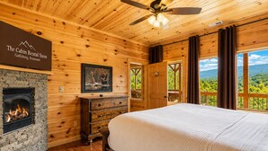A cabin rental store bedroom with a fireplace, wooden walls, a bed, a ceiling fan, and a view of the outdoors. - Sycamore Mansion | 14 Suites | Arcade | Fire Pit | Amazing Views (Gatlinburg)