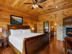A wooden cabin bedroom with a bed, a ceiling fan, a painting, a stone fireplace, and a bathroom visible through an open door.