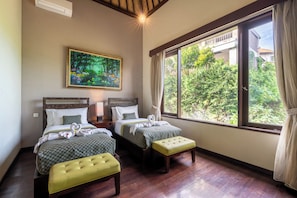 A hotel room with two beds, a painting, a window with a view of greenery, and a wooden ceiling.