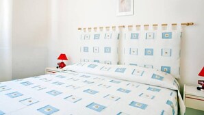 Een netjes opgemaakt bed met een bedsprei met patroon, een rode lamp op een nachtkastje, en een ingelijst beeld aan de muur.