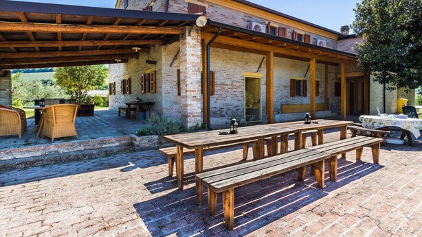 Une terrasse extérieure avec des tables et des bancs en bois, un bâtiment en briques surmonté d’une pergola en bois, et un ciel d’un bleu limpide.