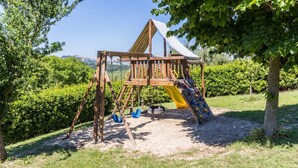 Une structure de jeux en bois, dotée d’un toboggan et de balançoires, entourée de verdure et d’arbres.