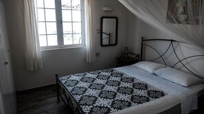 Un dormitorio con cama, ventana con cortinas, mesita de noche con lámpara y un espejo fijado a la pared.