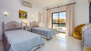 A hotel room with two beds, a large window with a view of a pool, a ceiling fan, and a small table with a lamp.