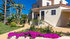 A two-story house with a tiled roof, a balcony with a railing, and a garden with purple flowers and palm trees.
