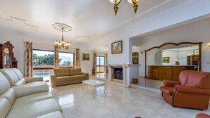 A spacious living room with a beige sofa, a brown leather armchair, and a white coffee table. A fireplace is visible in the background, and there is a large mirror on the wall.