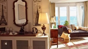 A living room with a sofa, a coffee table, and a lamp. There are posters on the wall and a window with a view of the sea. - Morlaco apartment (Málaga)