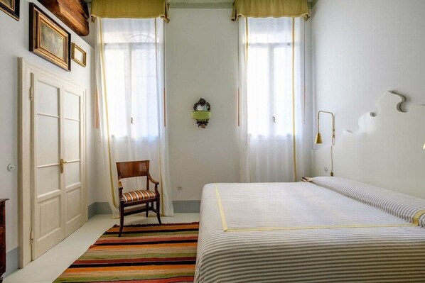 A bedroom with a bed, a chair, a window with curtains, a wall-mounted lamp, and a door. - Ca' del Ramo D'oro (Venice)