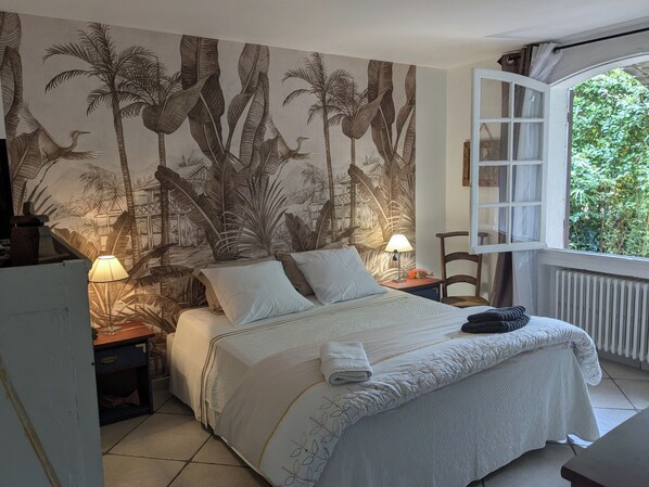 A bedroom with a bed, two bedside tables with lamps, a chair, and a window with a view of greenery.