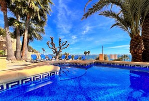 A swimming pool with blue mosaic tiles, surrounded by palm trees and lounge chairs.