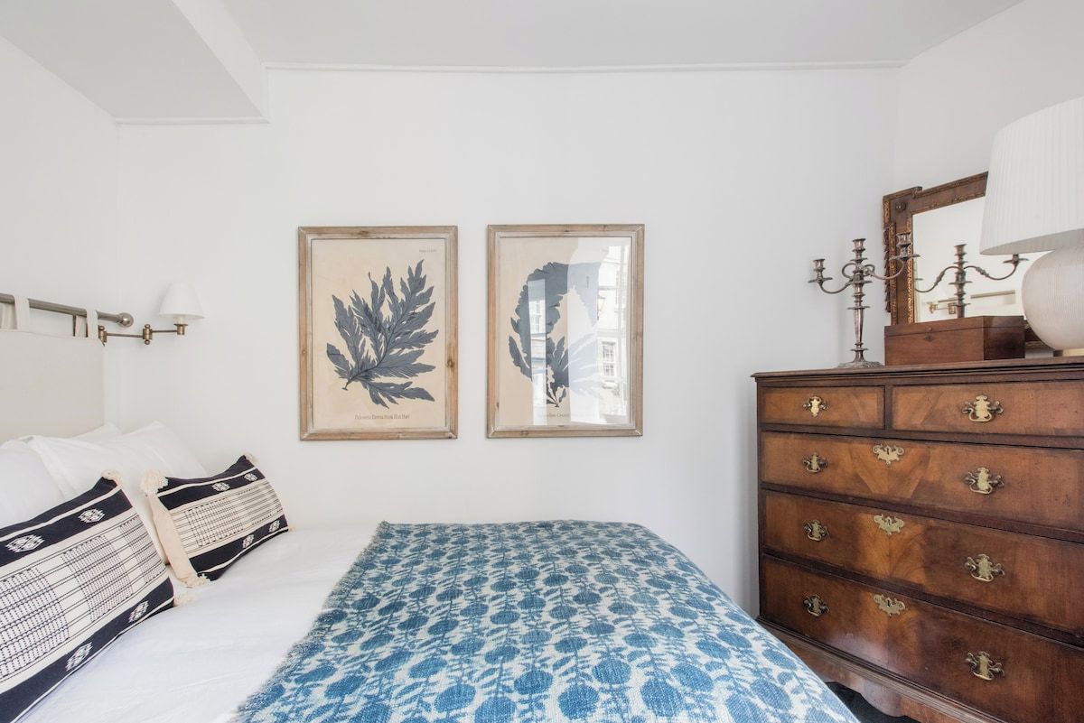 Quarto com cama, duas gravuras de plantas emolduradas, cômoda e mesa de cabeceira com abajur.