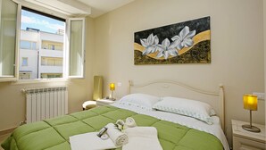 A hotel room with a green bedspread, a painting of flowers on the wall, a window with a view of buildings, and a bedside lamp. - Villa Mimosa Near Desenzano Old Town Apt 2 Parking, A/C, WiFi, Desenzano Del Garda, Italy (Desenzano Del Garda)