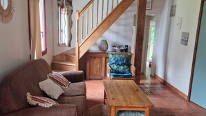 A spacious living room with a wooden staircase, a brown sofa with striped cushions, a wooden coffee table, and a blue patterned armchair. - Small house by the sea (Plouha)