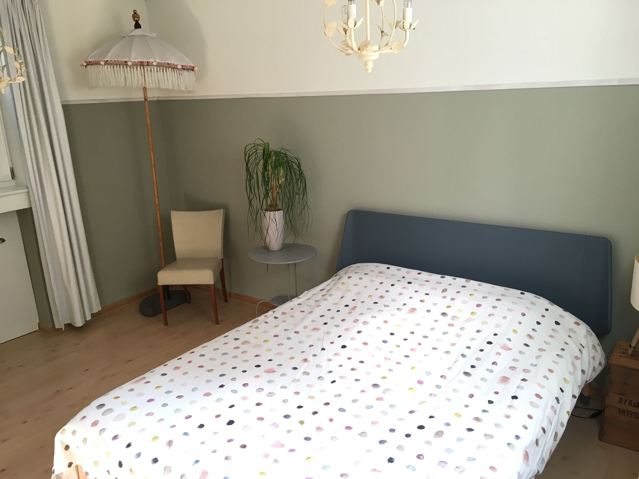 A bedroom with a bed, a chair, a small table with a plant, and a chandelier.