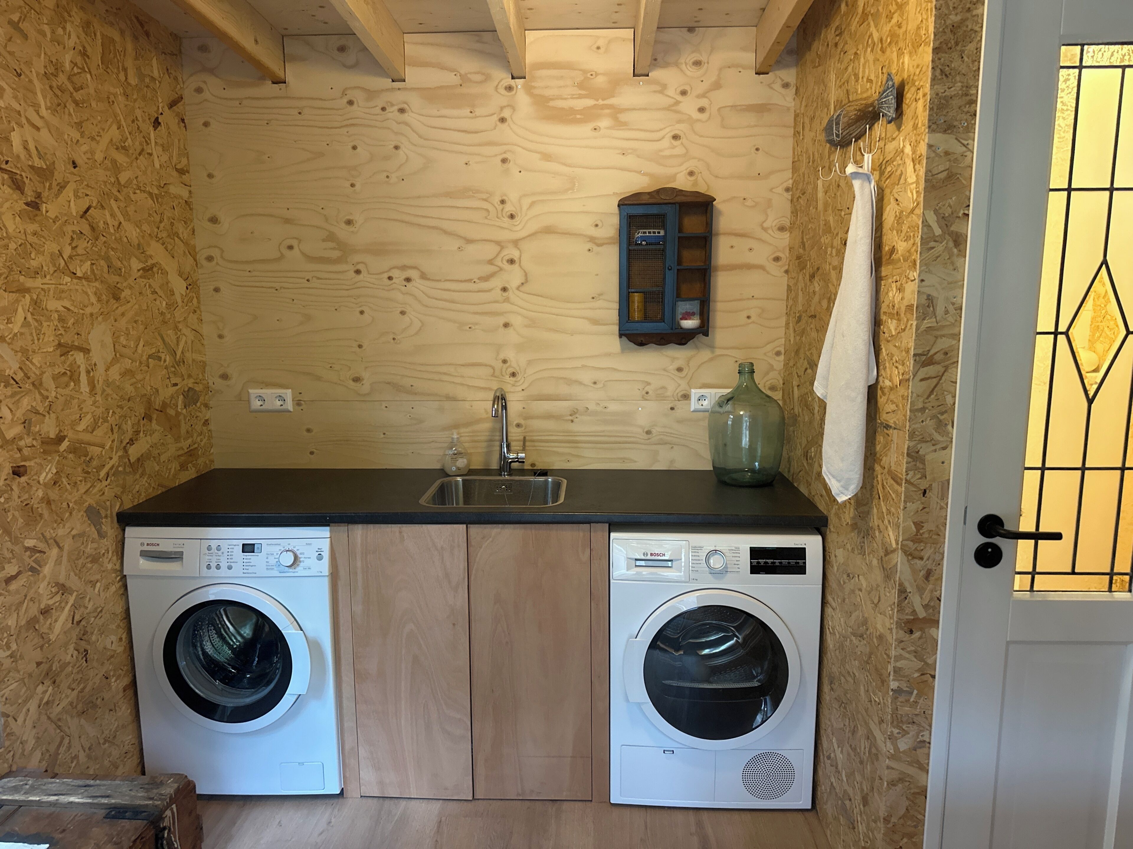 A compact laundry room with two Bosch washing machines, a sink, and a small shelf with a decorative item.