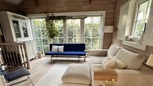 A cozy living room with a blue sofa, a beige sofa, a wooden coffee table with a vase of flowers, and a large window with a view of greenery.