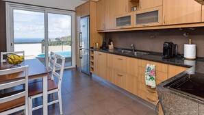 A kitchen with wooden cabinets, a stainless steel refrigerator, a black countertop, and a view of the sea through a sliding glass door. - 3-bedroom villa in Calheta, sea-view | Casa Belita (Calheta)