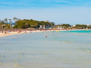 A beach with clear water, people, and buildings in the background.