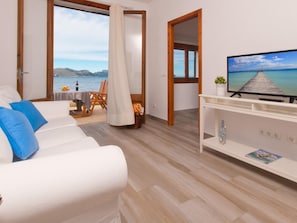 Un moderno salón con un sofá blanco, un televisor sobre un mueble blanco y vista al mar a través de un ventanal grande.
