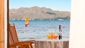 Ein Tisch mit einer Flasche und Gläsern, ein hölzerner Stuhl und der Blick auf eine Person beim Windsurfen auf dem Wasser mit Bergen im Hintergrund.