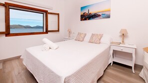 Een slaapkamer met een groot bed, een raam met uitzicht op zee, een schilderij aan de muur en een nachtkastje met een lamp.