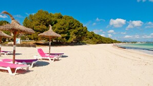 Ein Strand mit weißen Liegestühlen und Reetdachschirmen, ein klarer Himmel und ein bewaldeter Hügel im Hintergrund.