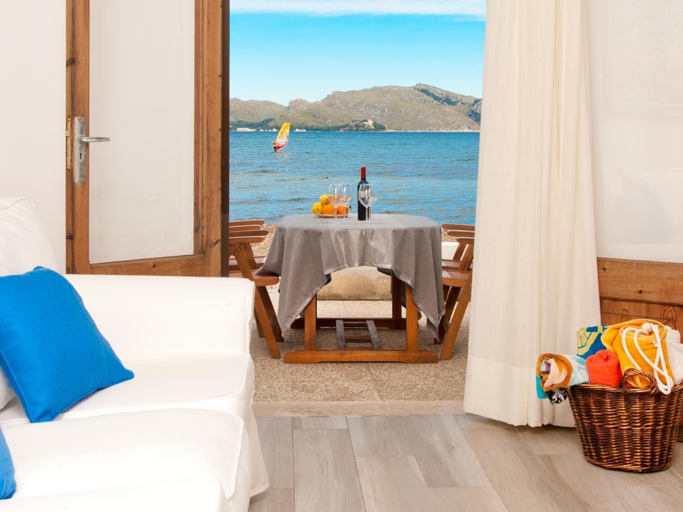A room with a bed, a table with a tablecloth, a wicker basket, and a view of the sea with a person windsurfing.