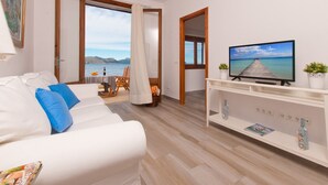 Ein modernes Wohnzimmer mit einem weißen Sofa, einem Fernseher auf einer weißen Konsole und Meerblick durch ein großes Fenster.