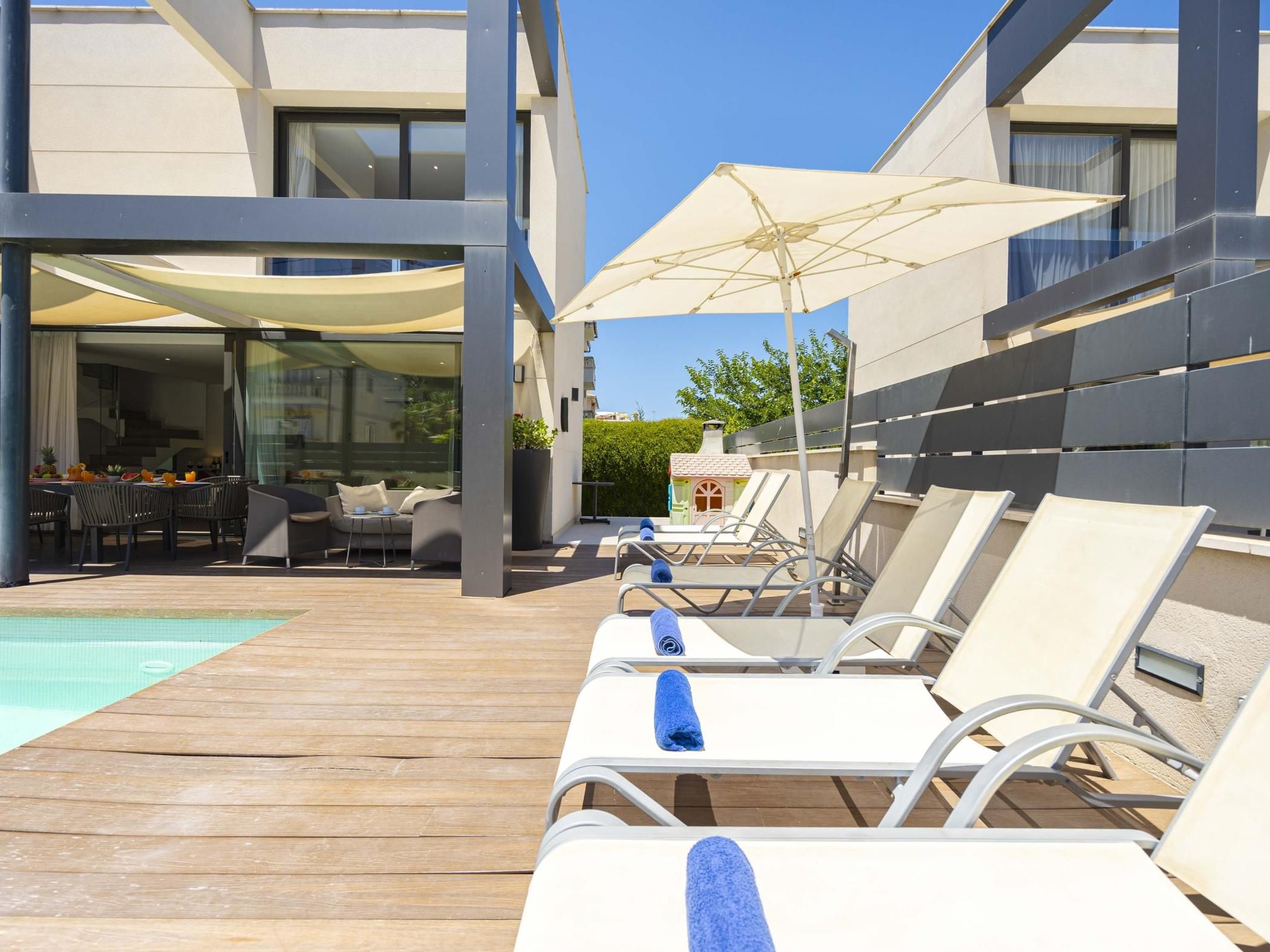 A poolside area with lounge chairs and a table, a wooden deck, and a building with large windows.