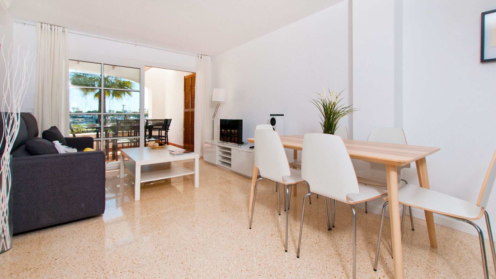 A modern living room with a sofa, a dining table with chairs, a TV, and a view of the outdoors through large windows.