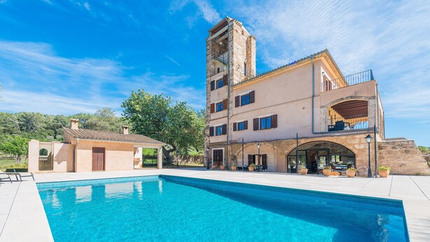 Vista de la piscina, un edifici, una piscina blava cristal·lina, una construcció petita a prop i una torre de pedra.