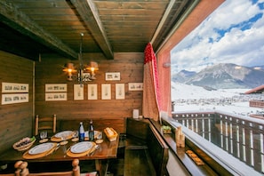 Un interior de madera con una mesa de comedor preparada para dos personas, cuadros en la pared y vistas a montañas nevadas.