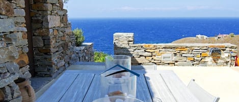 Ein Holztisch mit drei Glaskolben, gefüllt mit weißen Kieselsteinen und einem braunen Gegenstand, vor einer Steinsandsteinmauer und Blick aufs Meer.