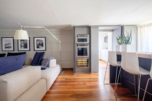 A modern living room with a sofa, a dining area with chairs, a built-in oven, and framed pictures on the wall.