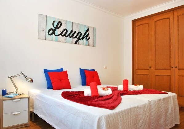 A neatly made bed with red and blue pillows, a red blanket, and a 'Laugh' sign on the wall.