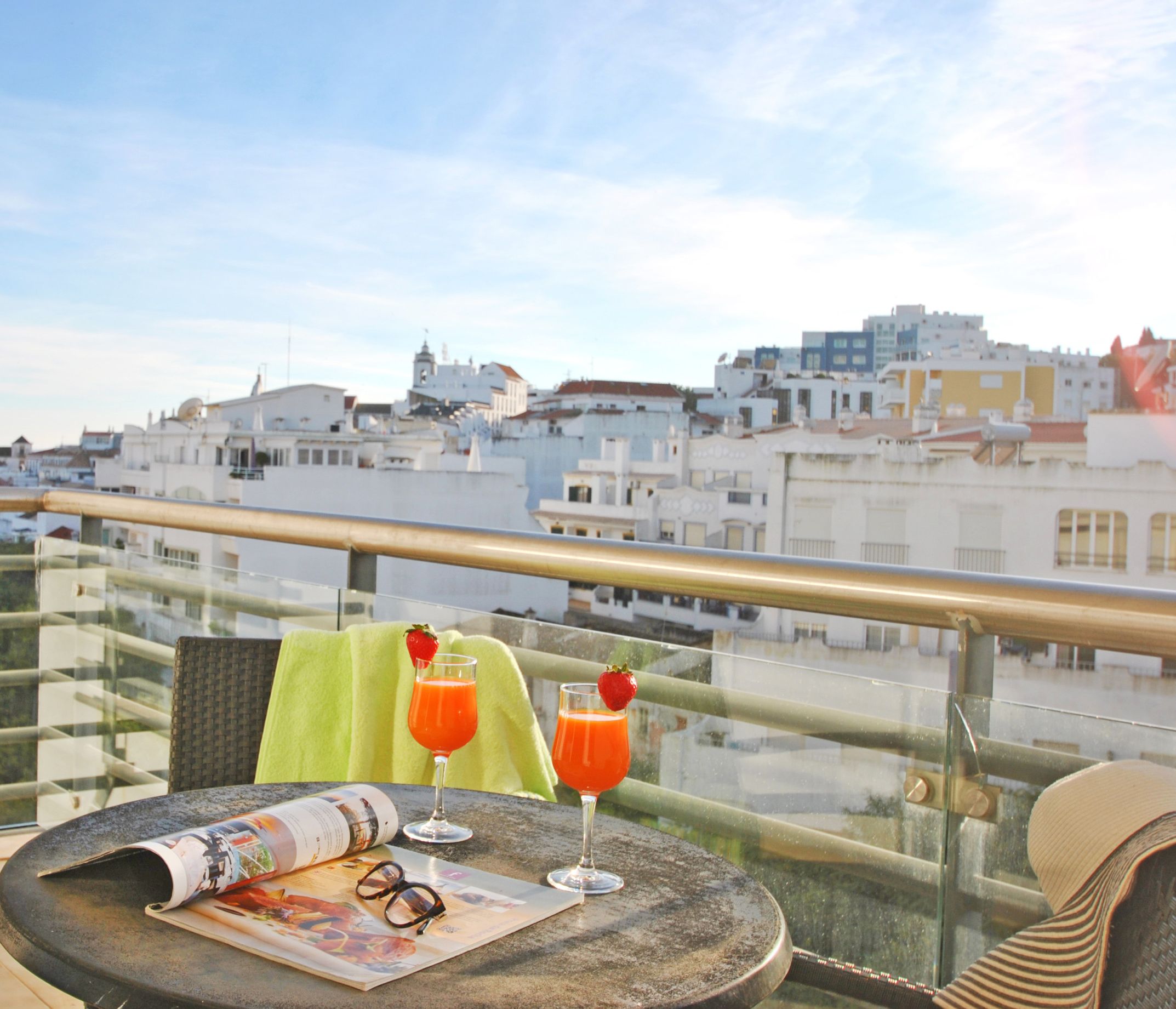 Un balcon avec une table dressée pour deux personnes, agrémenté d’un magazine, d’une paire de lunettes et de deux boissons. Le balcon offre une vue sur un paysage urbain avec des bâtiments blancs.