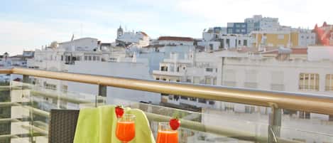 Un balcón con una mesa preparada para dos personas, con una revista, un par de gafas y dos bebidas. El balcón ofrece vistas a un paisaje urbano con edificios blancos.