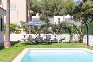 Una piscina con sillas de descanso y una sombrilla, rodeada por una valla blanca y árboles.