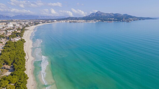 Vue aérienne d’une zone côtière avec une plage, des eaux d’un bleu limpide et un paysage montagneux en arrière-plan.