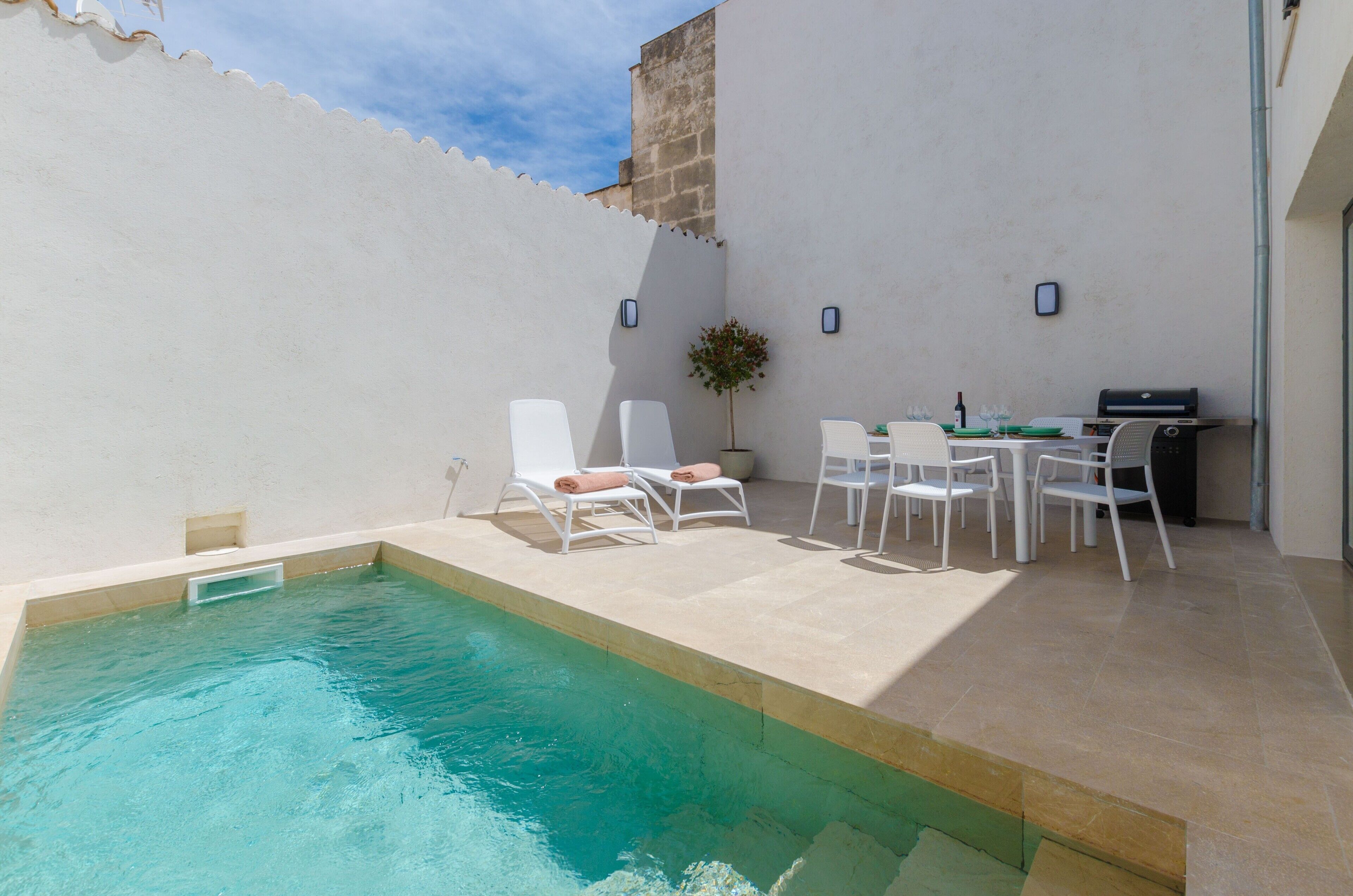 Una piscina con sillas de descanso y una pequeña mesa con sillas, rodeada por un muro blanco y un edificio de piedra.