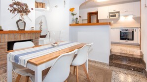 A dining area with a wooden table and white chairs, a brick fireplace, and a kitchen in the background.
