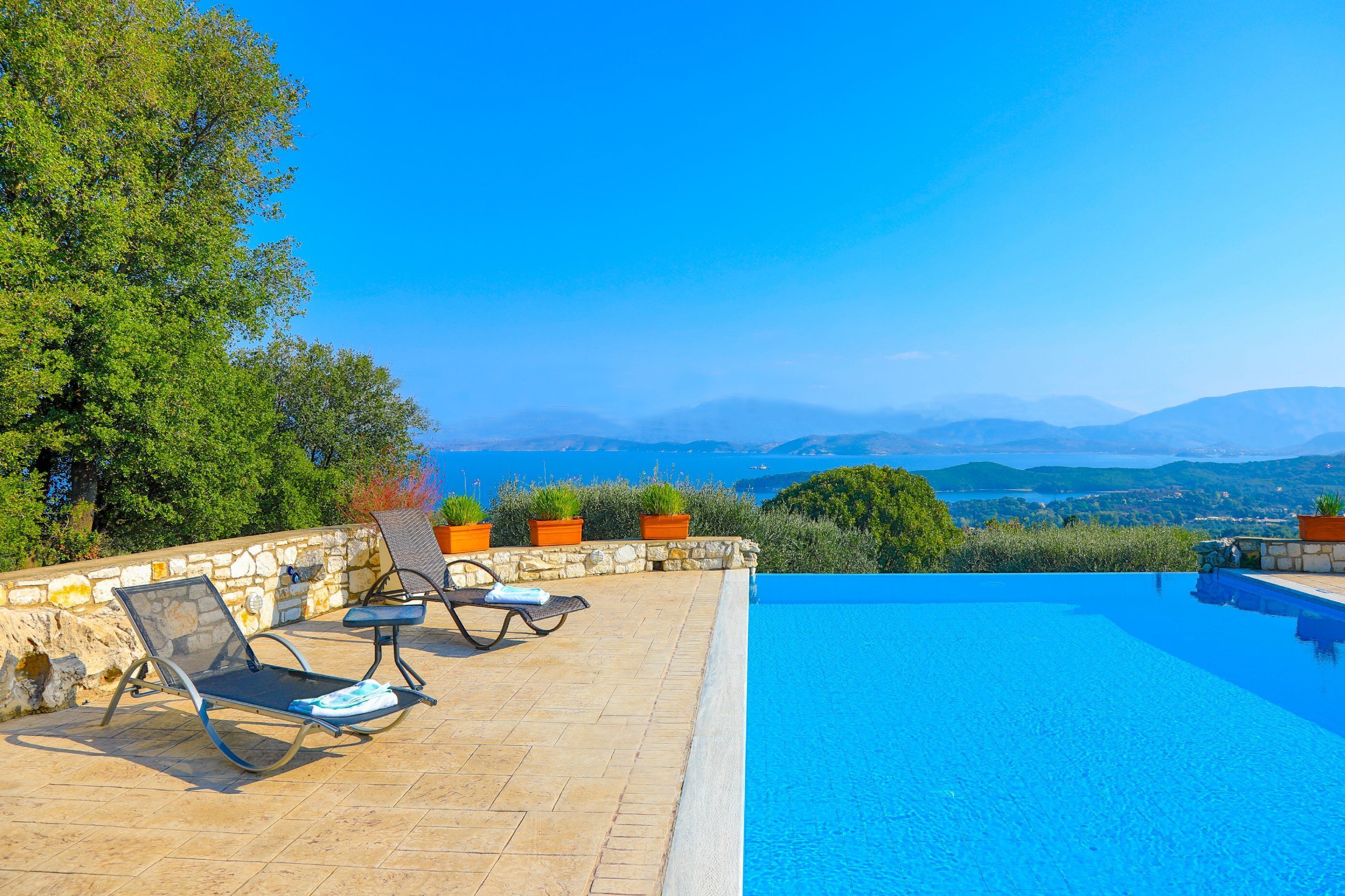 Un espace aménagé au bord de la piscine, avec des chaises longues et un mur en pierre, offrant une vue panoramique sur un paysage magnifique, avec des montagnes et un ciel d’un bleu éclatant.
