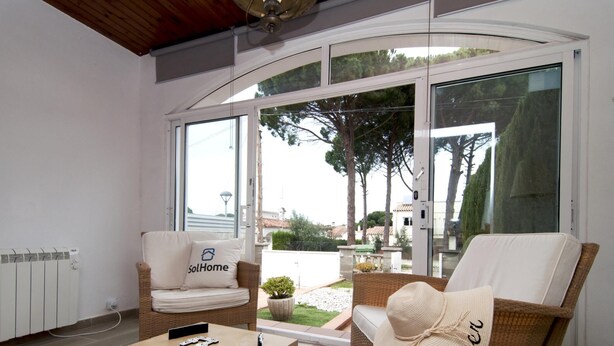 Una sala de estar moderna con mobiliario de mimbre, una mesa de centro de madera y vistas a un jardín y edificios a través de amplios ventanales.