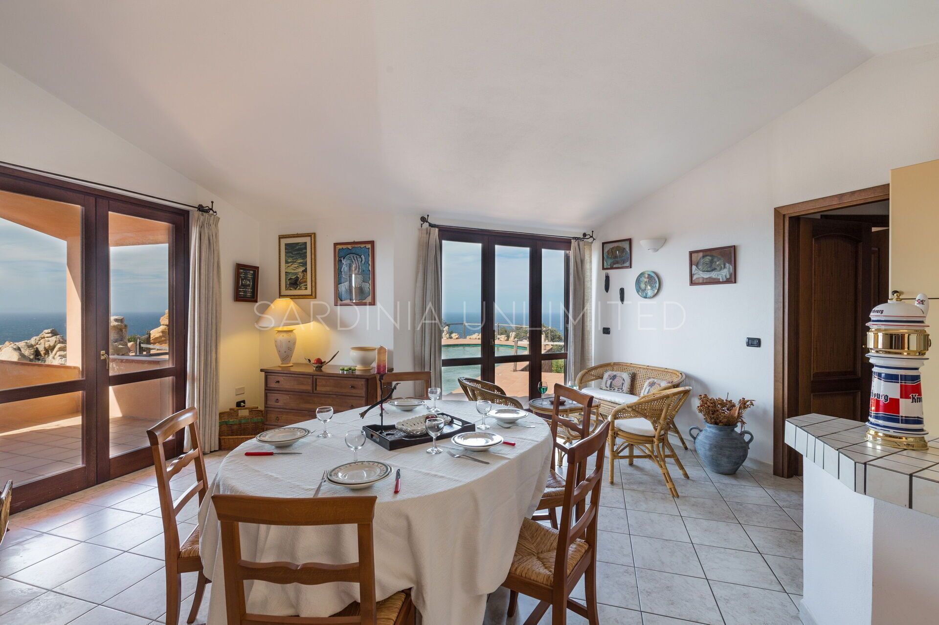 Un comedor con una mesa redonda dispuesta para cuatro personas, rodeada de sillas de mimbre. Se aprecia vista al mar a través de puertas de vidrio amplias.